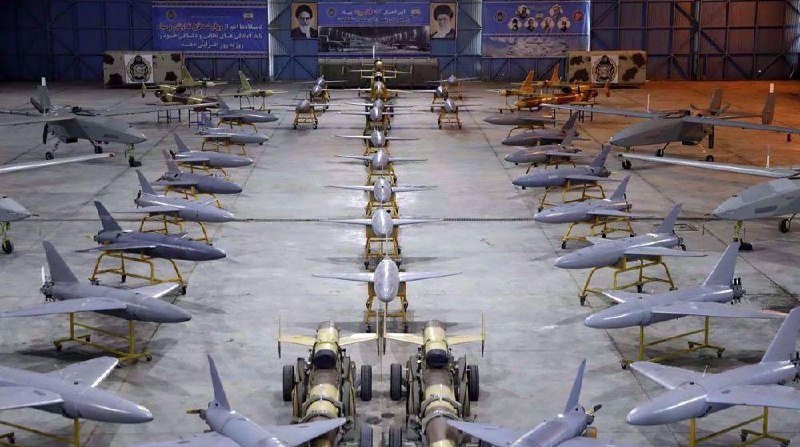 Rows of unmanned aerial vehicles and missiles are arranged in formation inside a hangar, displayed in front of banners bearing portraits and Persian text.