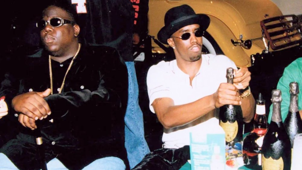 Two men wearing sunglasses sit at a table with several champagne bottles, one opening a bottle.