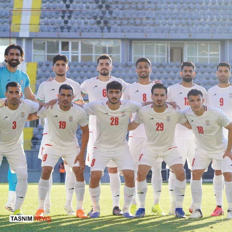 Iran national football team players in white kits pose for a team photo on a stadium pitch with empty blue seats in the background.
