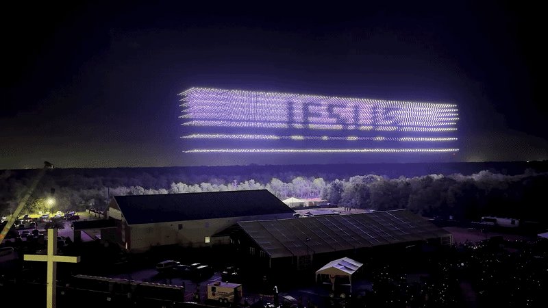 Hundreds of illuminated drones form the word "JESUS" in the night sky above a rural property with barns, vehicles, and a lit cross.