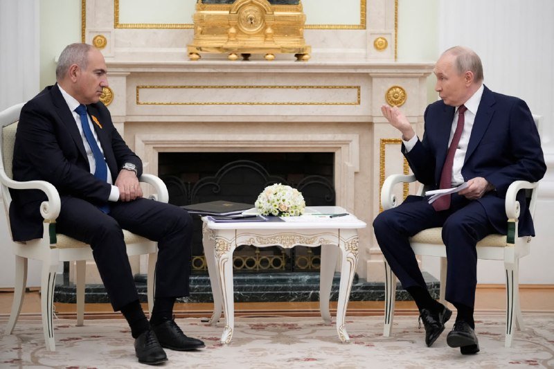 Two men in dark suits sit in ornate chairs across from each other in a formal room with a marble fireplace, one gesturing while speaking as the other listens.