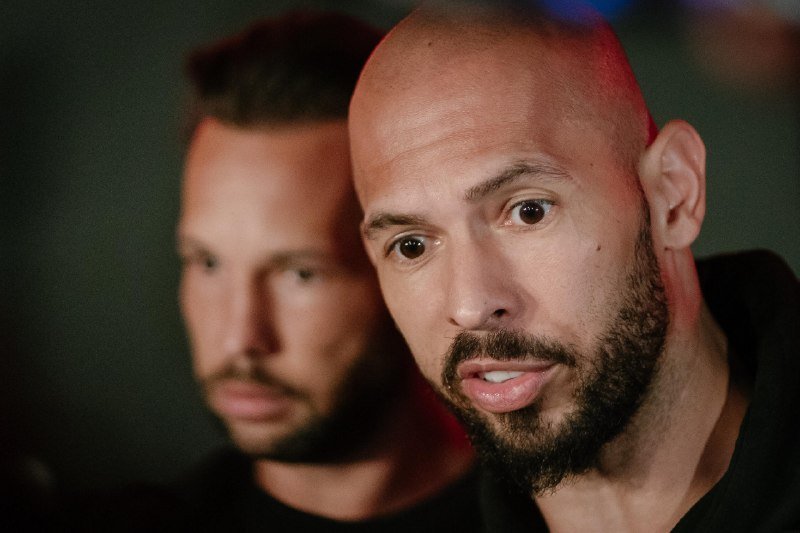 Two bearded men with closely cropped hair sit side by side against a dark background, with the man in the foreground looking directly at the camera with an open-mouthed expression.