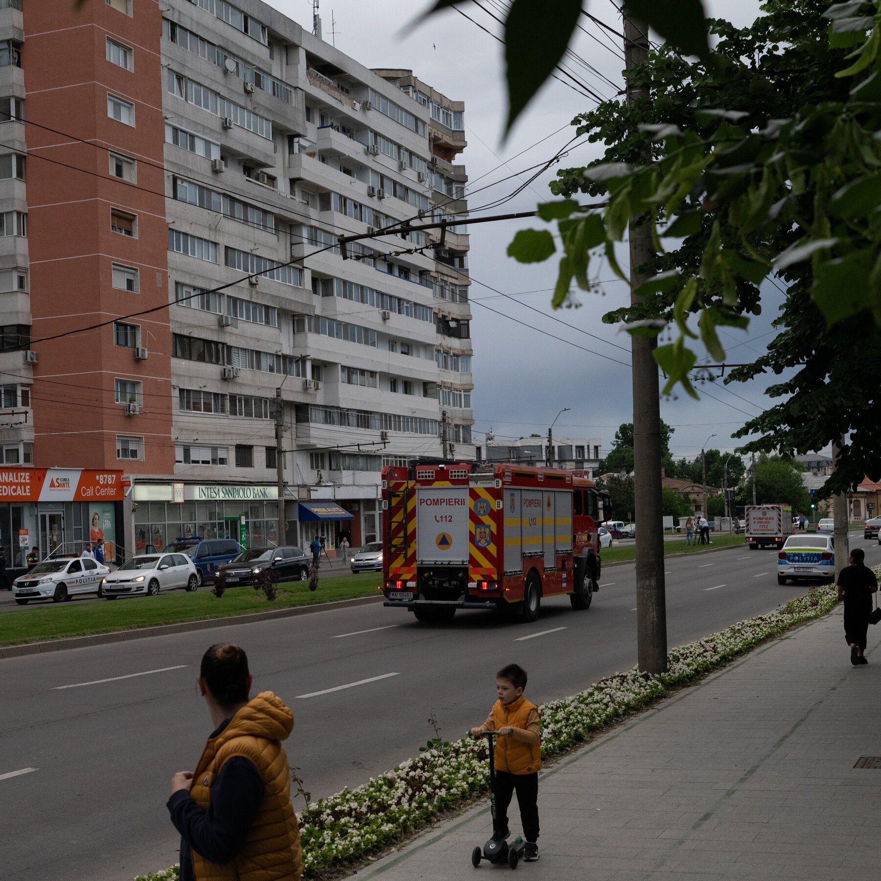 A red fire truck marked "POMPIERII" drives down a city street past parked police cars, a bank, and pedestrians on a sidewalk.