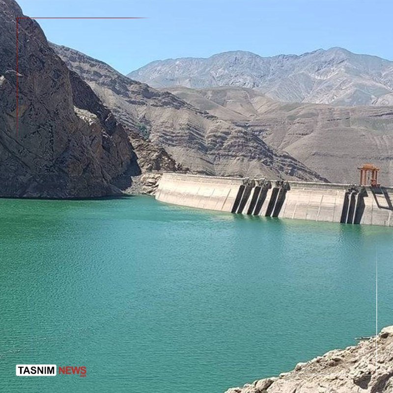 A concrete dam holds a large turquoise reservoir, with rugged, arid mountains rising under a clear sky in the background; a "TASNIM NEWS" watermark appears in the corner.