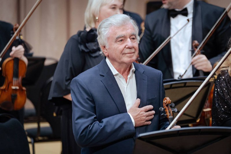 A gray-haired man in a dark suit stands with his hand on his chest, surrounded by formally dressed musicians holding violins and bows on stage.