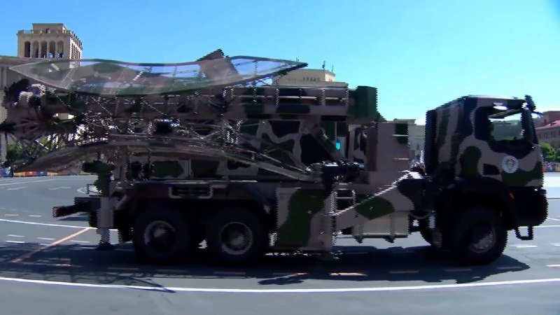 A camouflaged military truck carrying a large radar or antenna system drives along a paved road during a daytime parade.