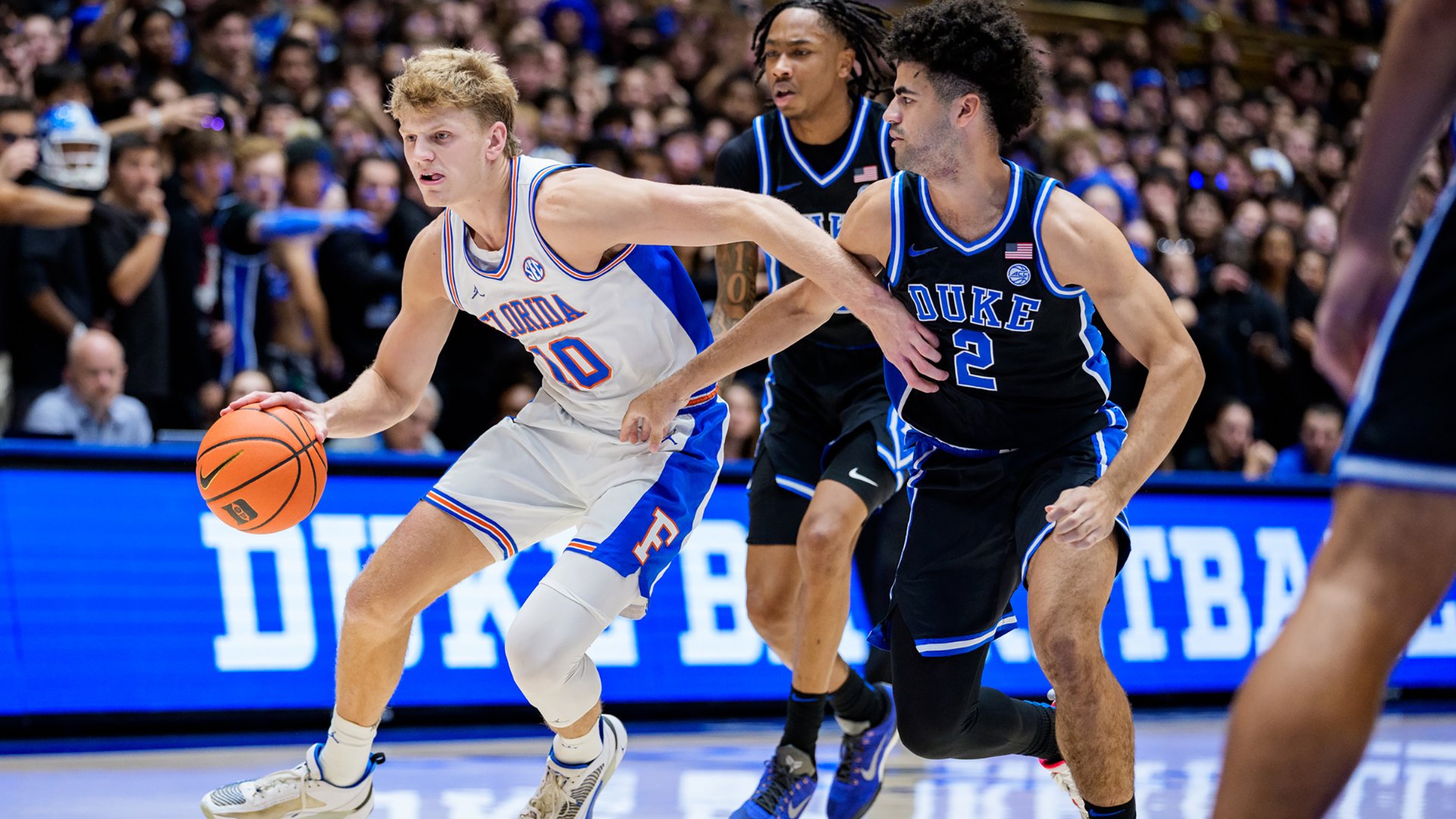 A Florida basketball player in a white jersey dribbles against two Duke defenders in black jerseys during a college basketball game in front of a crowded arena.