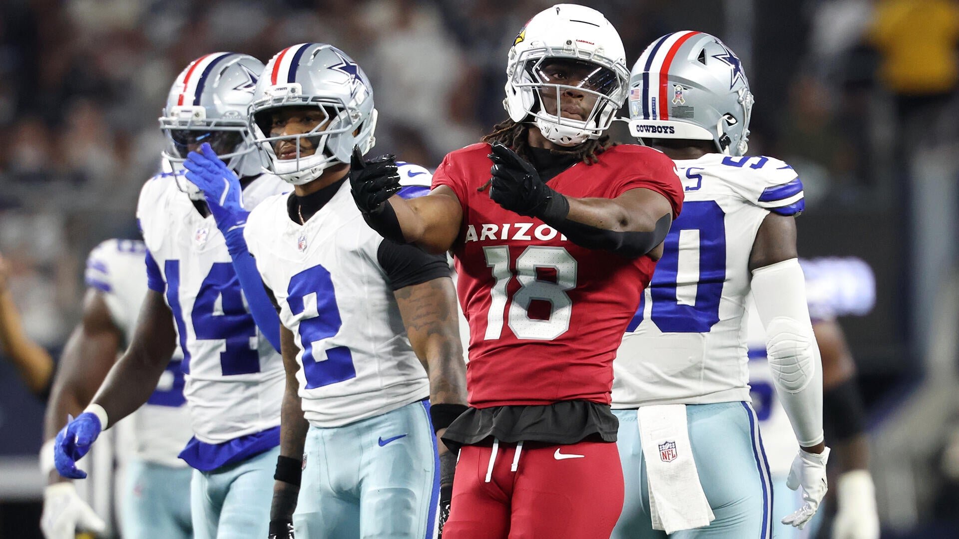 An Arizona Cardinals player wearing jersey number 18 gestures with his hand while surrounded by three Dallas Cowboys defenders in white uniforms during an NFL game.