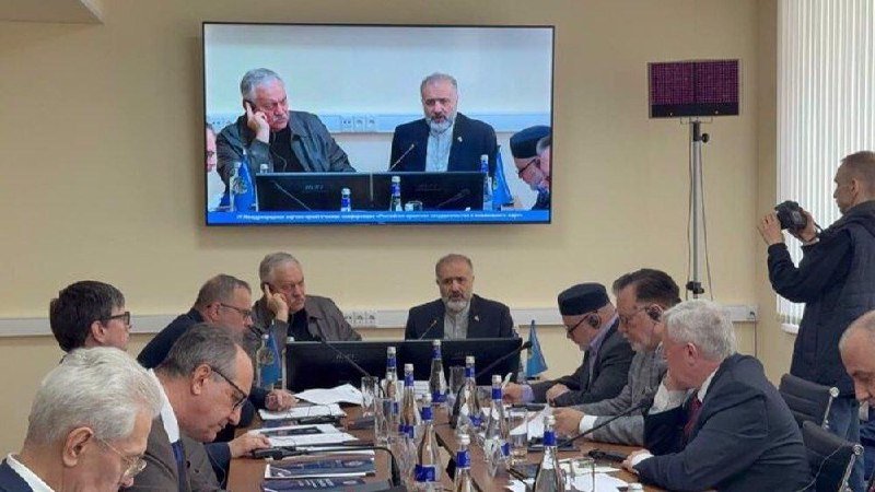 A group of men sit around a conference table with water bottles and documents, while a large wall-mounted screen displays two other men seated at a similar meeting, and a photographer takes pictures.