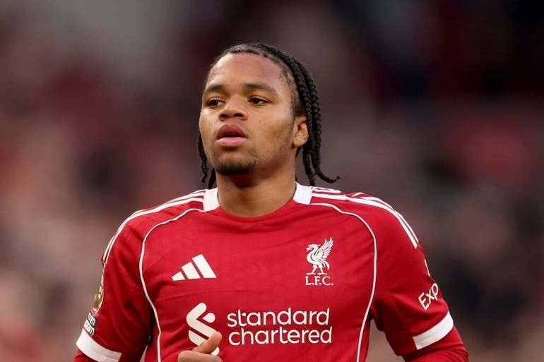 A soccer player with braided hair wearing a red Liverpool FC jersey with "Standard Chartered" sponsorship looks on during a match.
