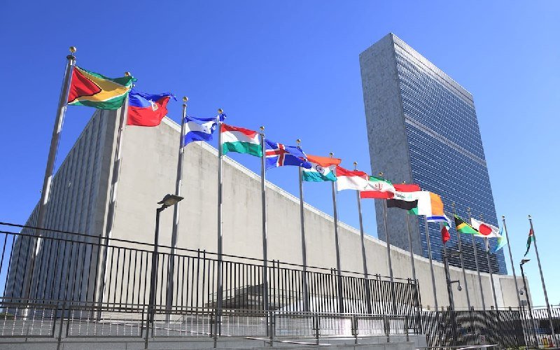 A row of international flags flies on flagpoles in front of a large modernist concrete building and a tall glass skyscraper under a clear blue sky.