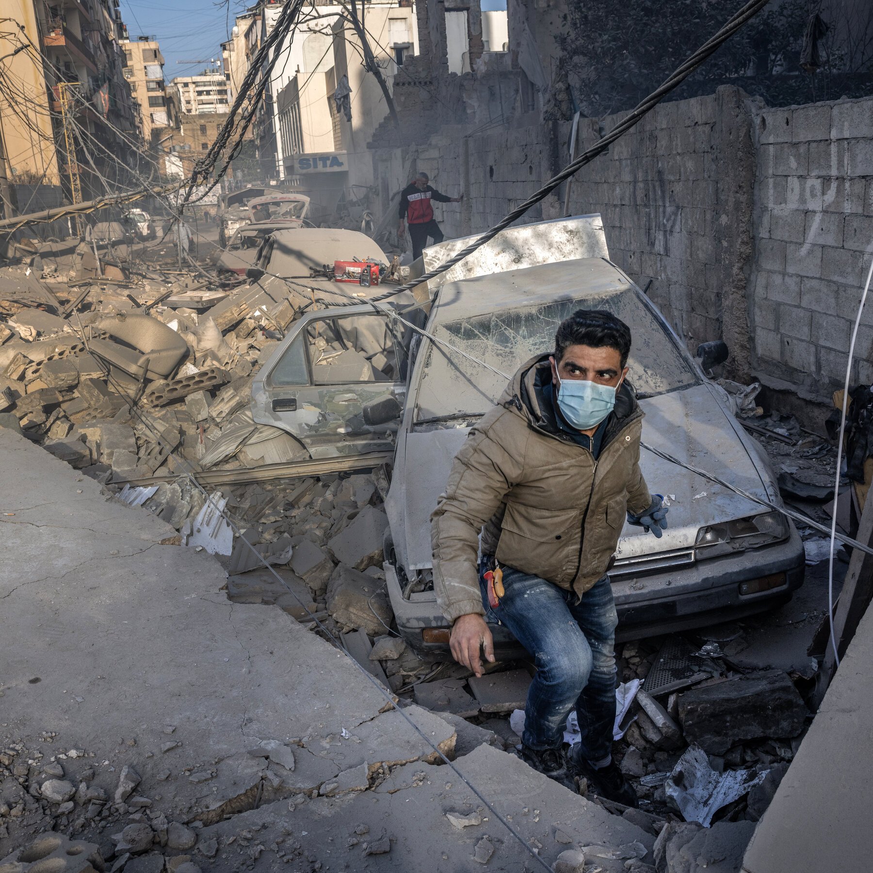 A masked man walks through a debris-filled street with crushed cars, tangled wires, and damaged buildings visible in the background.