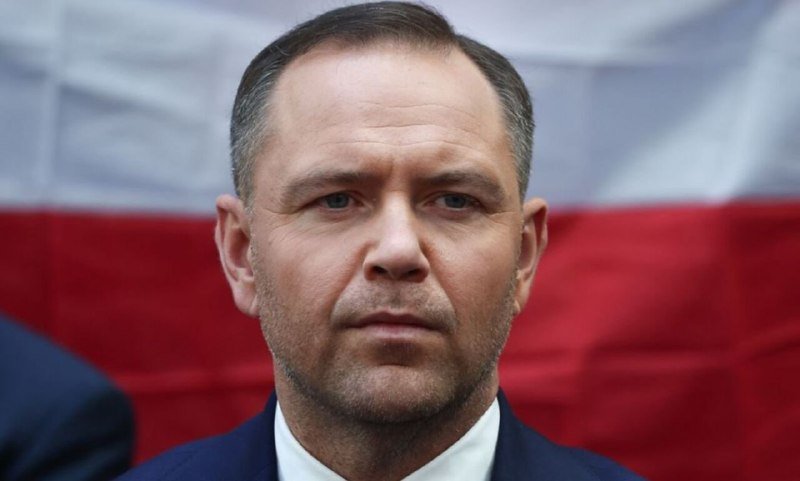 A man in a dark suit and white shirt stands before a white and red flag, looking off-camera with a serious expression.