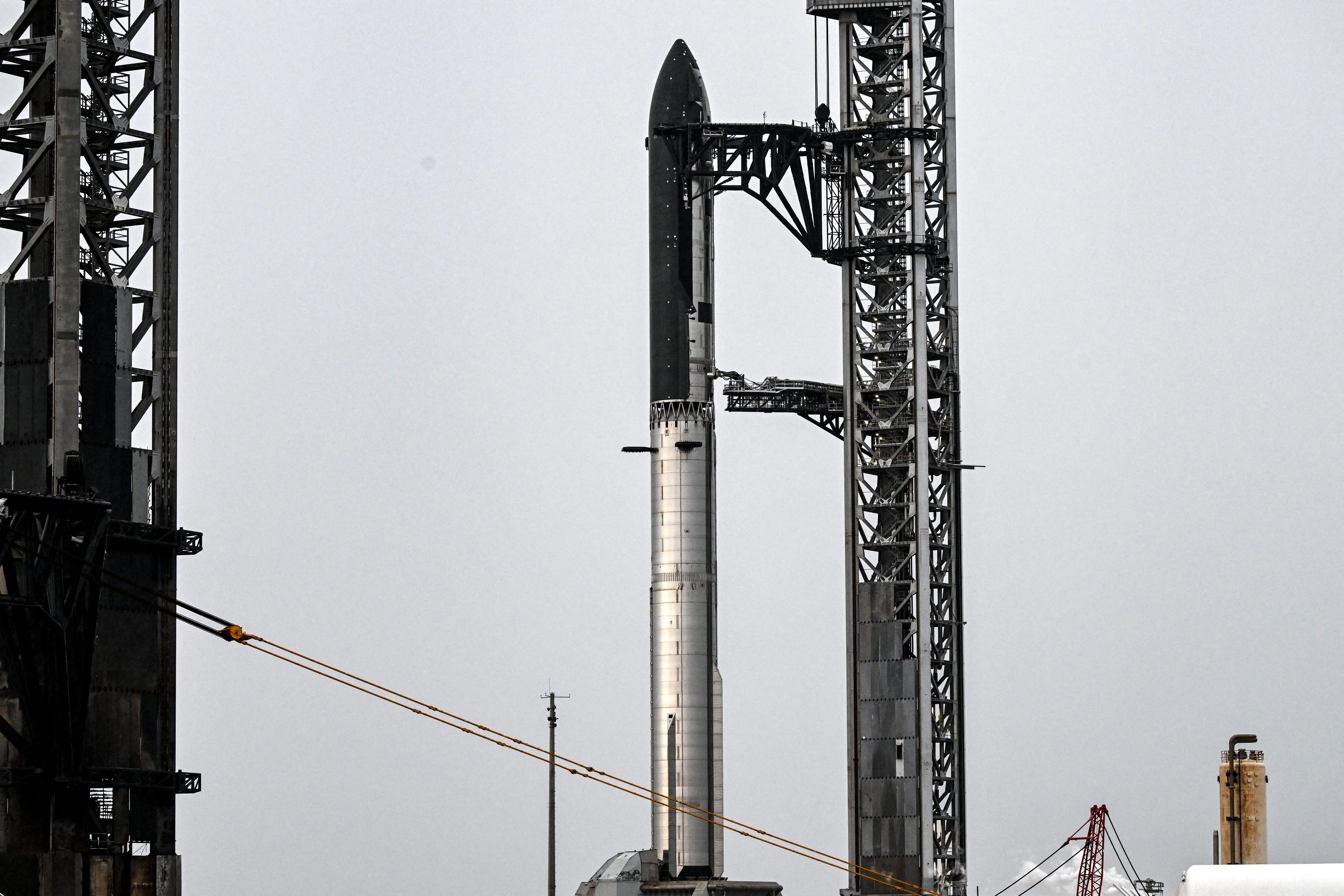 A rocket lifts off from a snowy launch pad, flames and thick smoke billowing as it ascends past a nearby launch tower and support structures.