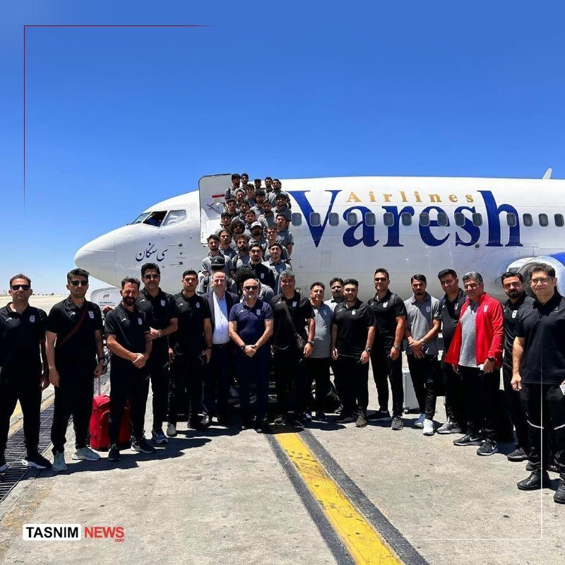 A large group of men pose for a photo on a tarmac in front of a white "Varesh Airlines" passenger jet under a clear blue sky.