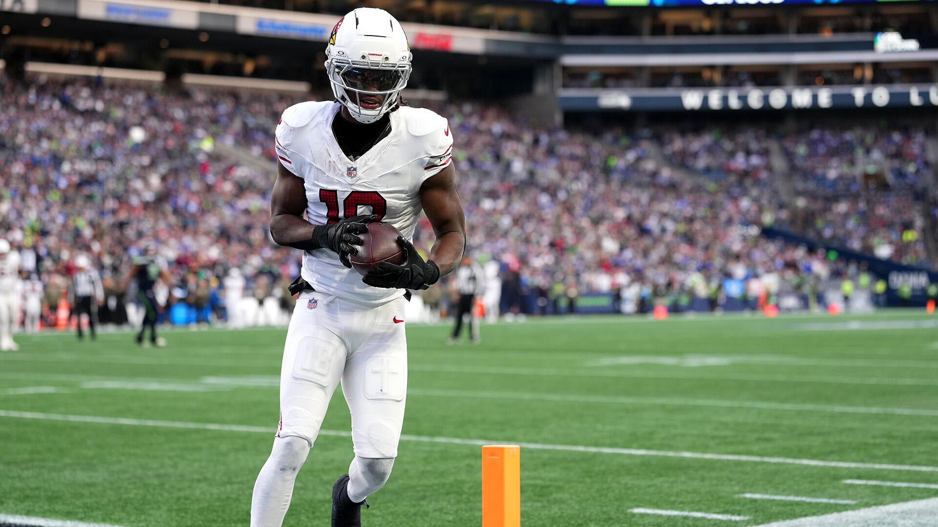 An Arizona Cardinals player wearing a white uniform and helmet runs with the football in the end zone of a packed stadium.