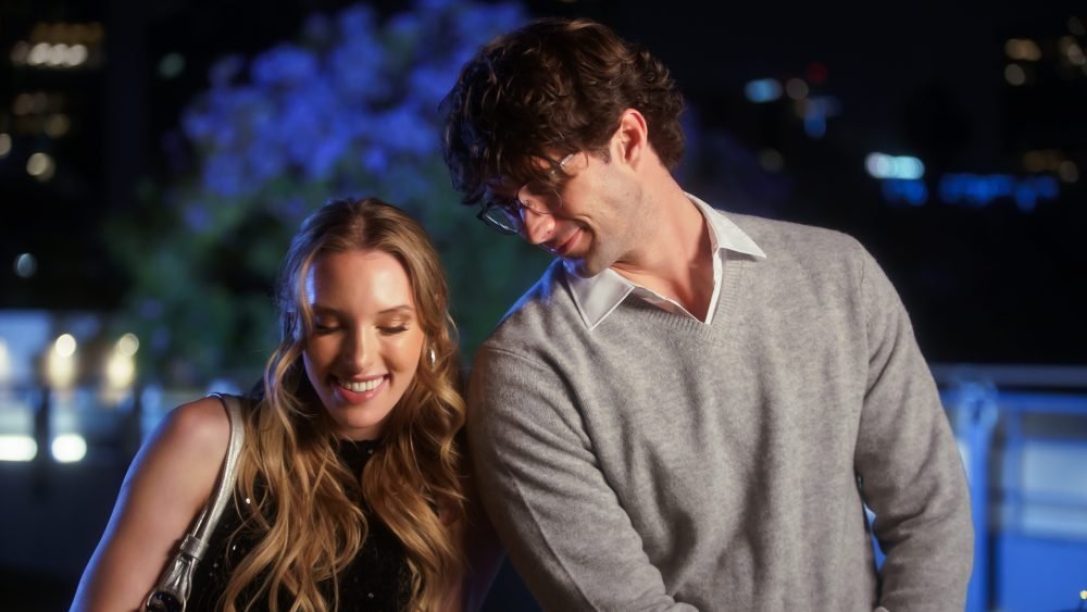 A smiling woman with long blonde hair stands beside a smiling man wearing glasses and a gray sweater at night, with blurred city lights in the background.