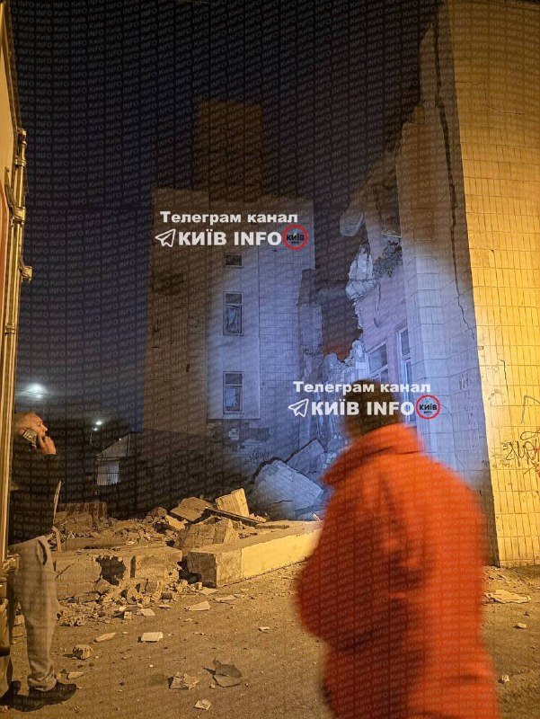A nighttime scene shows a partially collapsed multi-story building with rubble on the ground, as two figures in the foreground observe the damage.