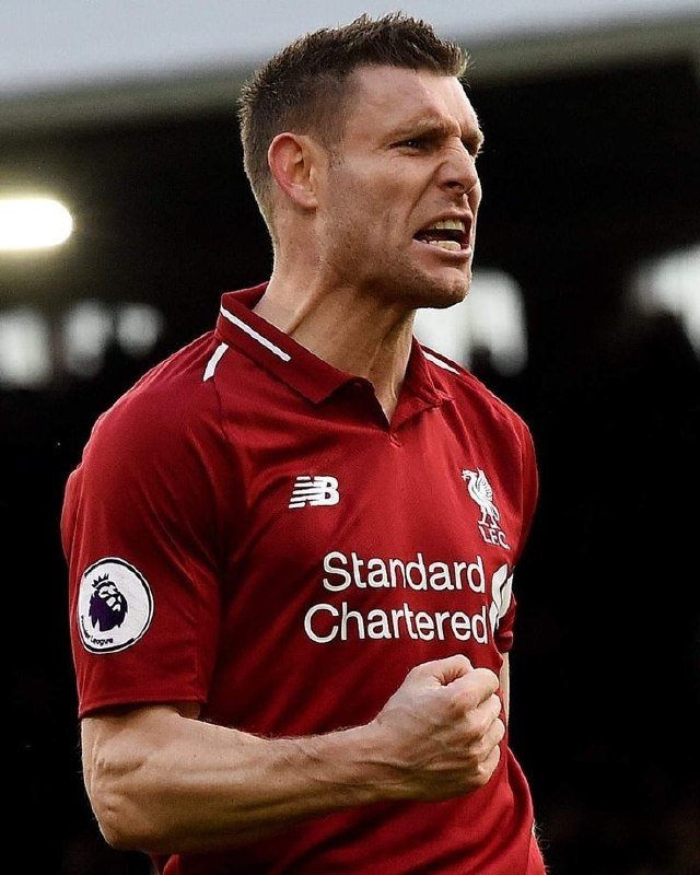A man with short brown hair, wearing a red Liverpool LFC soccer jersey featuring "Standard Chartered" and a Premier League patch, clenches his fist with an intense expression.