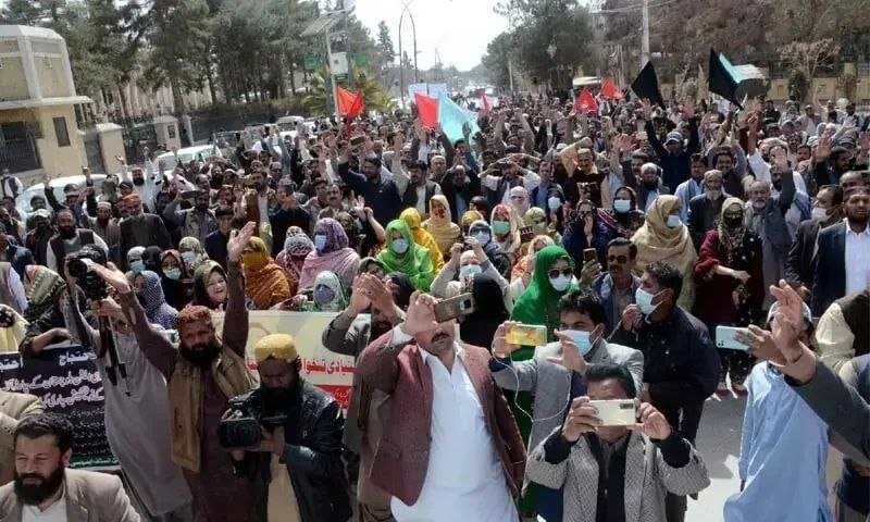A large crowd of people, many wearing face masks and holding up smartphones, march down a tree-lined street while waving black, red, and green flags.