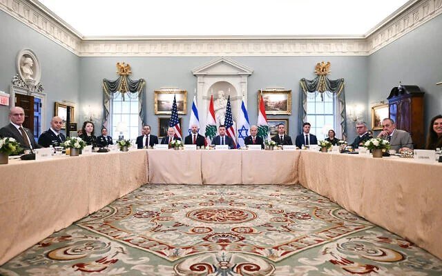 A large group of people in formal attire sit around a U-shaped conference table in an ornate, blue-walled room, with multiple national flags displayed in the background.