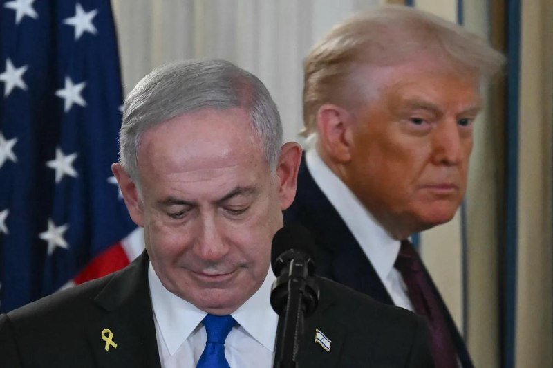 Two men in dark suits stand at a microphone with an American flag in the background, one looking down with a yellow ribbon pin on his lapel.