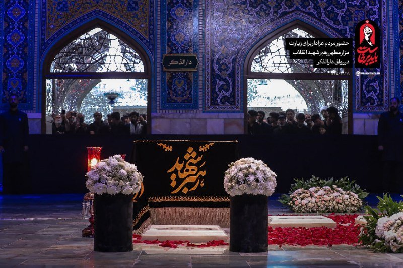 A black-draped shrine adorned with gold Arabic calligraphy stands flanked by white floral arrangements and scattered rose petals, with crowds of men gathered behind arched, mirrored doorways illuminated in blue light.