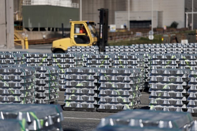 Rows of stacked silver metal ingots sit in an industrial yard, with a yellow forklift parked in the background near a building.