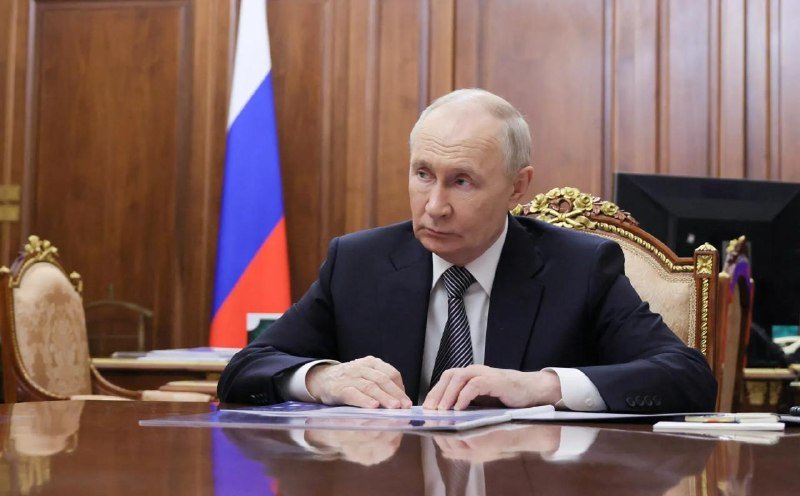 A man in a dark suit and striped tie sits at a polished wooden desk with documents, with a Russian flag visible behind him against wood-paneled walls.