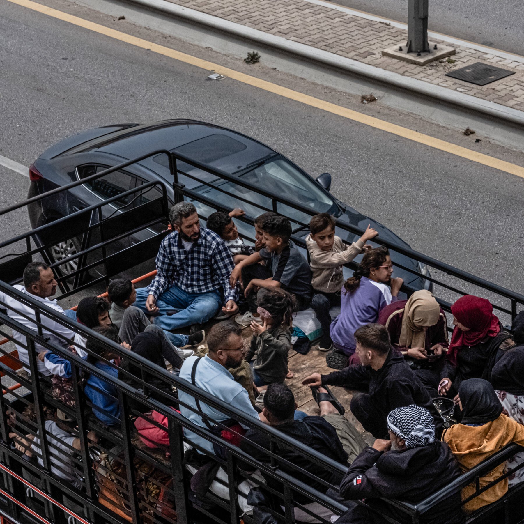 A crowded truck bed filled with men, women, and children sits on a paved road beside a black sedan.