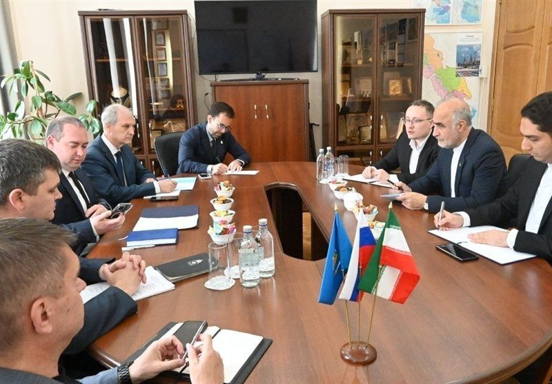 Several men in suits sit around an oval wooden conference table with water bottles, fruit, notepads, and small flags, taking notes in an office setting.