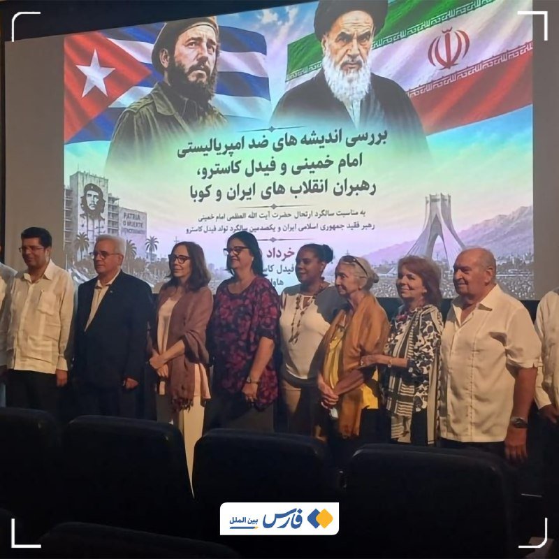 A group of people stand together in a theater before a large screen displaying Cuban and Iranian flags, portraits of two men, and Persian text.
