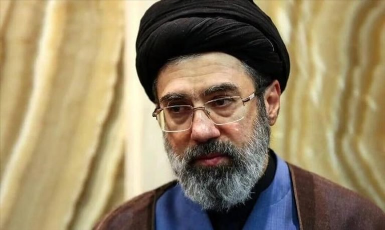 A bearded man wearing glasses, a black turban, and clerical robes stands before gold draped curtains.