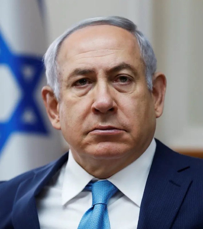 A gray-haired man in a dark suit and blue tie sits in front of a flag featuring a blue Star of David.