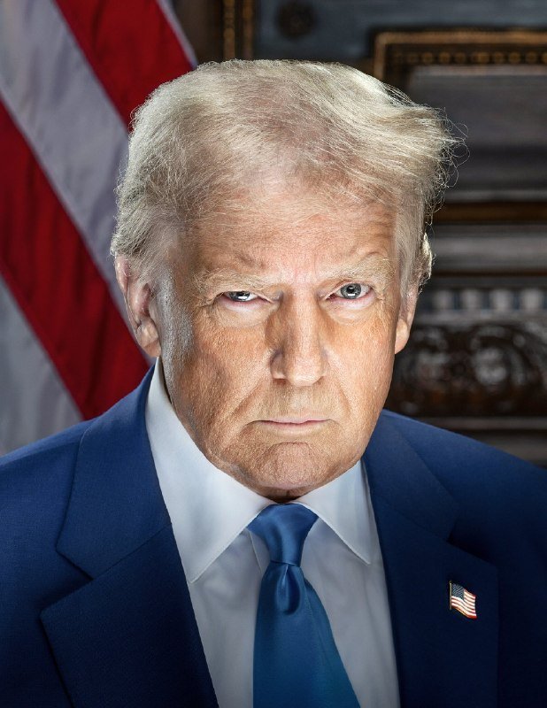 A man with blonde hair wearing a navy blue suit, white shirt, and blue tie with a U.S. flag pin looks sternly at the camera, with an American flag visible behind him.