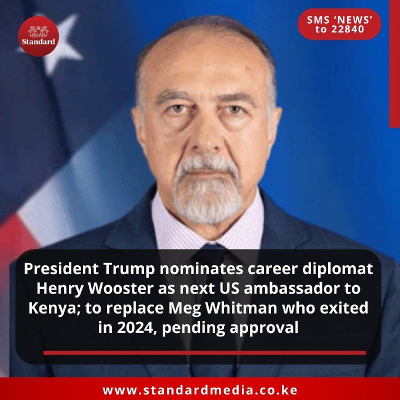 A bearded man in a suit and tie stands before an American flag, with a news caption about a diplomatic nomination.
