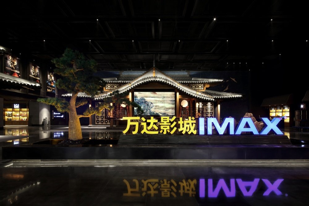 A traditional Chinese-style pavilion with upturned eaves is displayed indoors, flanked by a pine tree and illuminated "万达影城 IMAX" signage reflecting on the polished floor.