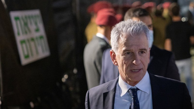 A gray-haired man in a dark suit and blue tie walks past a vehicle bearing Hebrew text, with people in red berets visible in the background.
