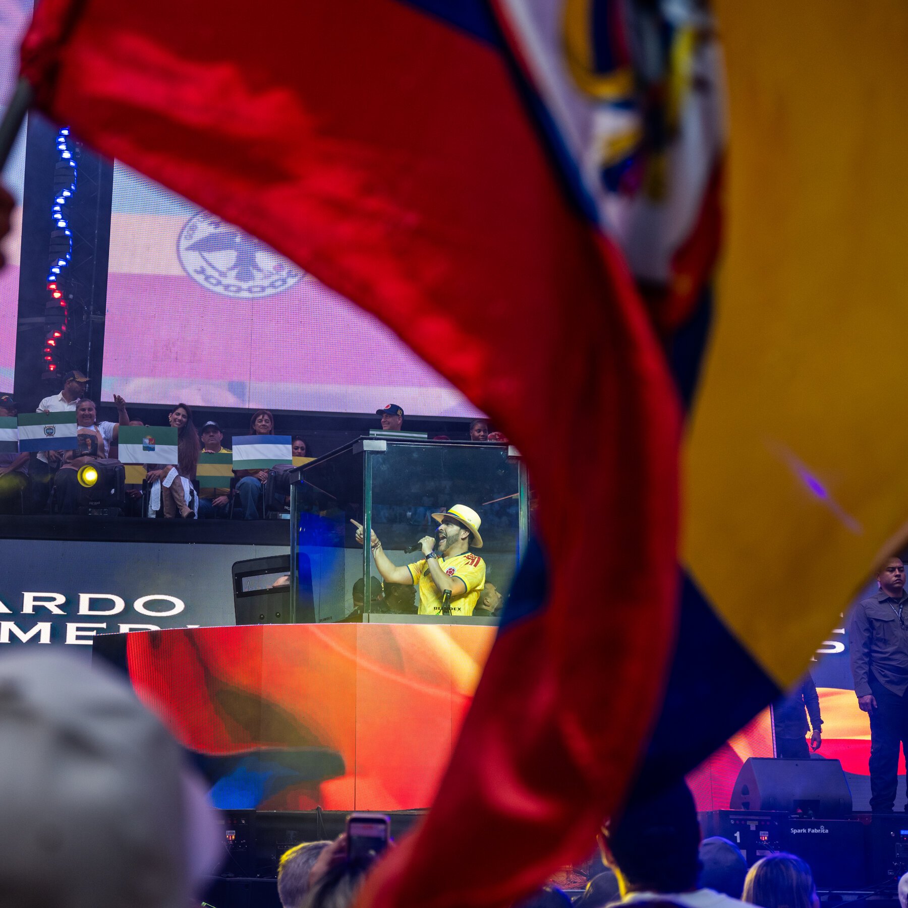 A man in a yellow Colombia jersey and hat speaks into a microphone inside a glass booth at a rally, with waving red and yellow flags in the foreground.