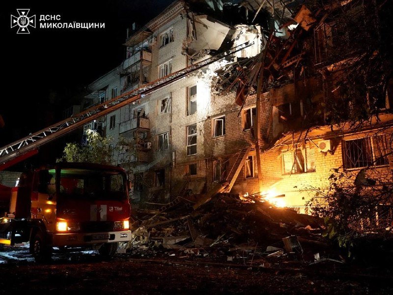A fire truck with an extended ladder is positioned at a heavily damaged multi-story brick apartment building at night, with debris scattered across the ground and flames visible amid the wreckage.