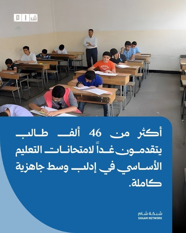 Students sit at wooden desks writing on papers in a classroom while a man in a white shirt stands observing them.