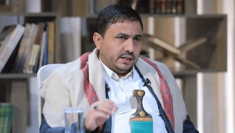 A man wearing a white shirt, dark vest, and red-and-white scarf speaks while seated in front of a bookshelf, with a water bottle and glass nearby.