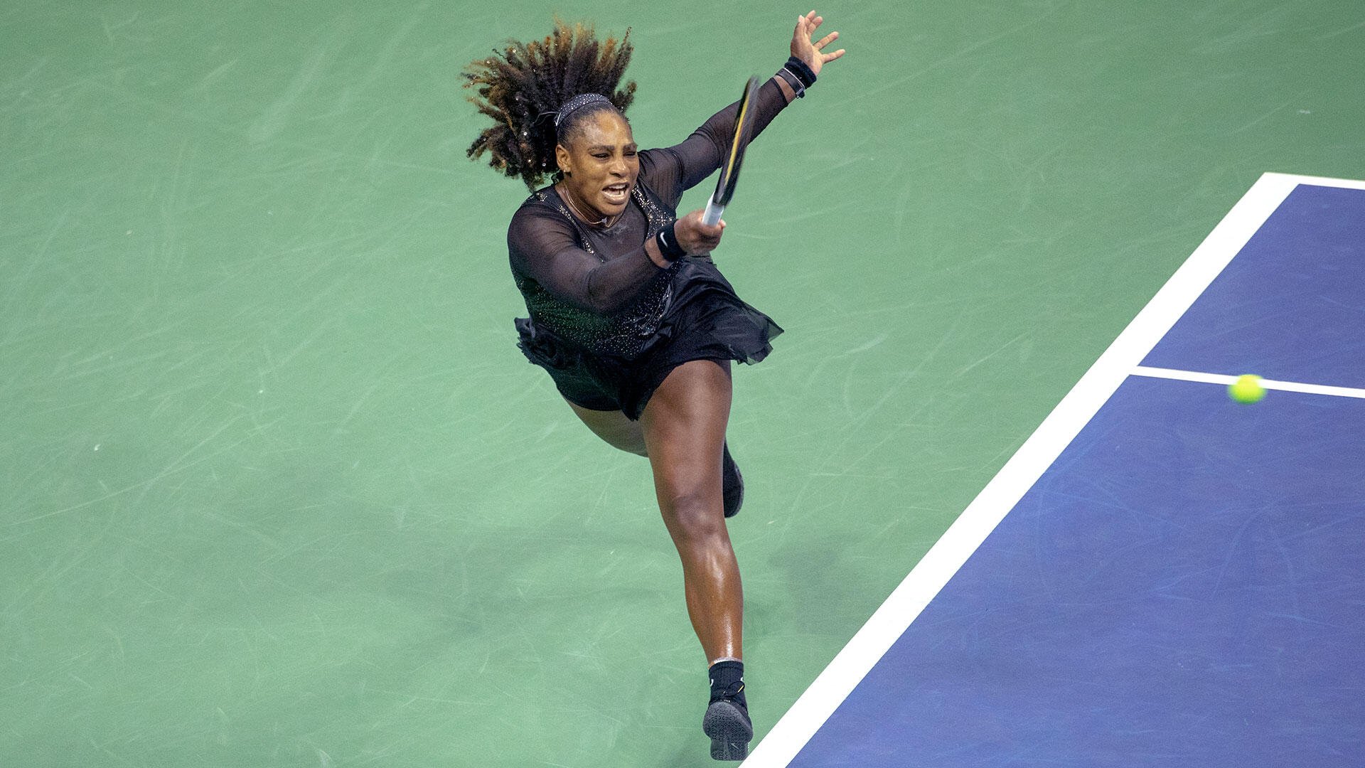 A female tennis player in a black dress swings her racket at a yellow ball on a green and blue hard court.