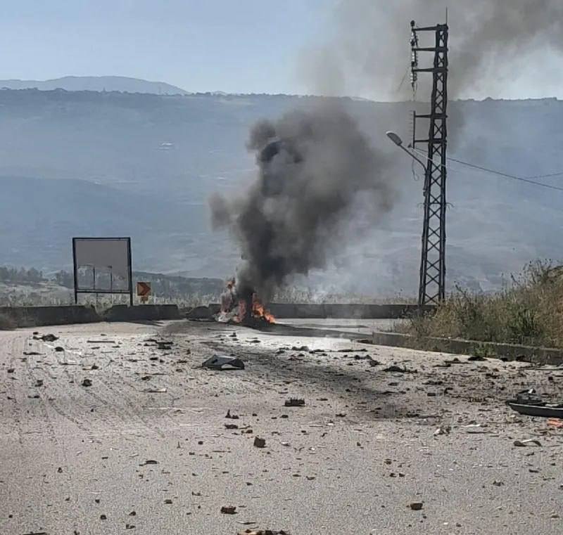 A vehicle burns on a rural road, sending a thick plume of black smoke into the sky amid scattered debris, with a metal power tower and hazy hills in the background.