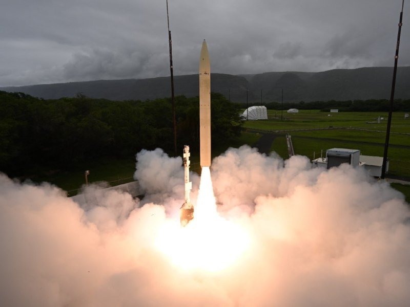 A rocket lifts off from a coastal launch pad, its bright exhaust plume billowing amid a cloudy sky and green fields.
