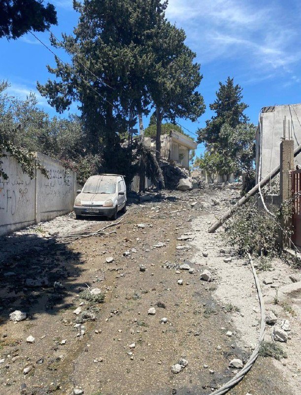A white van sits amid rubble and debris on a dirt road, with damaged structures, fallen utility lines, and trees under a blue sky.