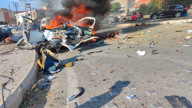A burning, destroyed white car sits on a street scattered with debris, with other damaged vehicles visible in the background amid heavy black smoke.