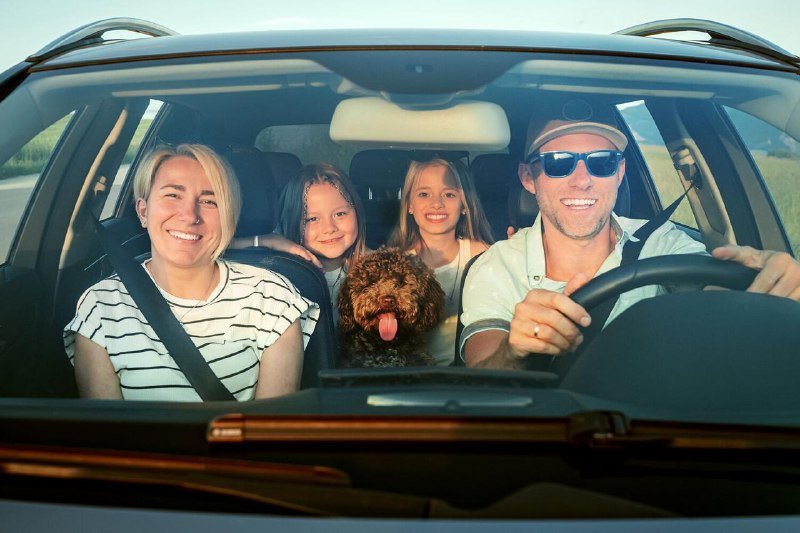 A family of four—two adults and two children—smile from inside a car alongside a small brown dog, with a man wearing sunglasses behind the steering wheel.