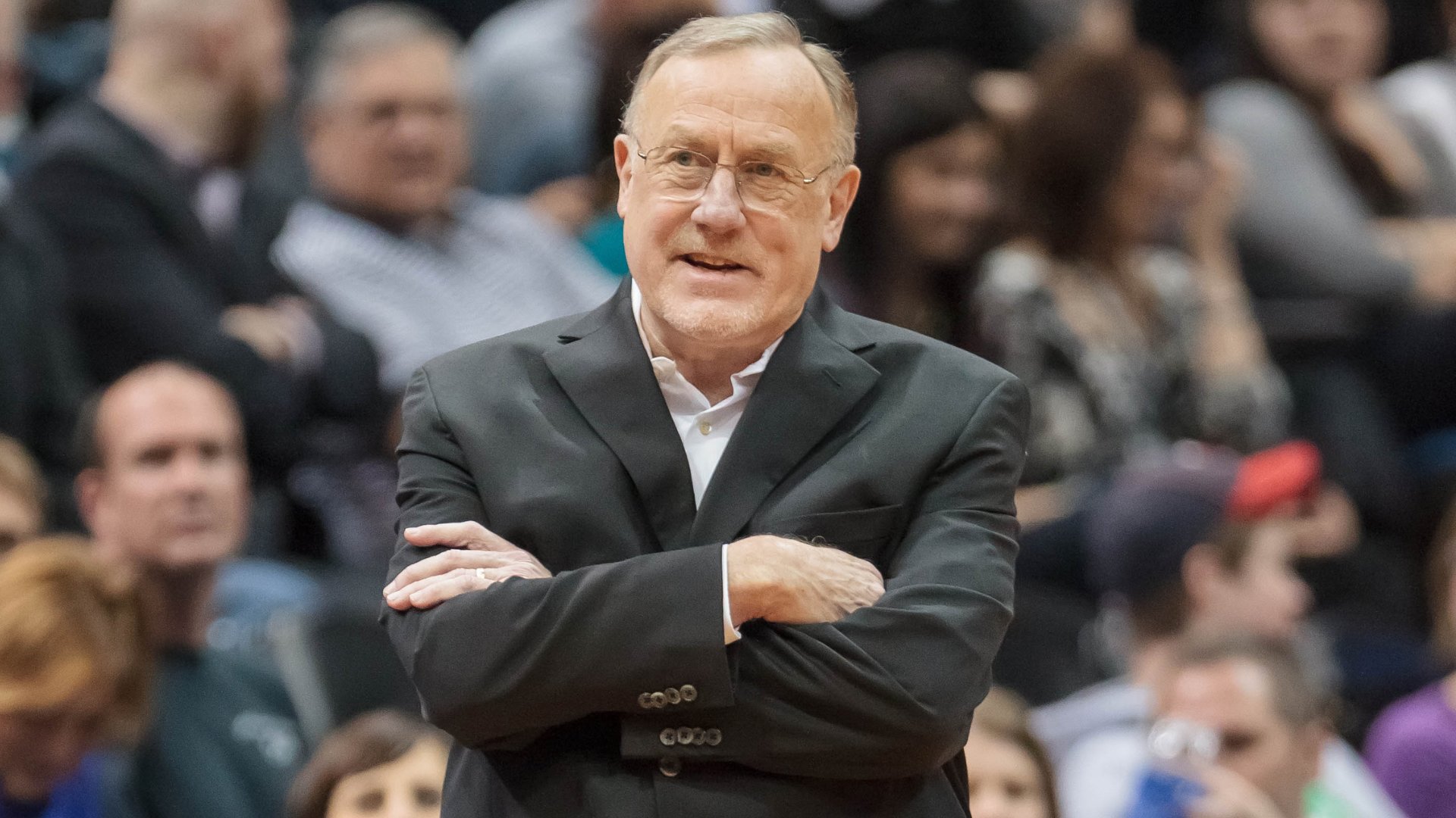 An older man with glasses, wearing a black suit and white shirt, stands with his arms crossed in front of a crowd of seated spectators.
