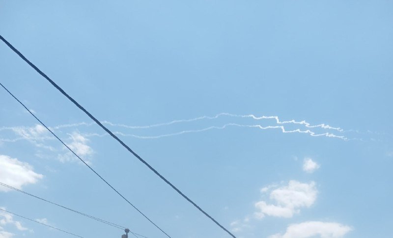 White smoke trails streak across a blue sky dotted with clouds, framed by dark power lines crossing the foreground.